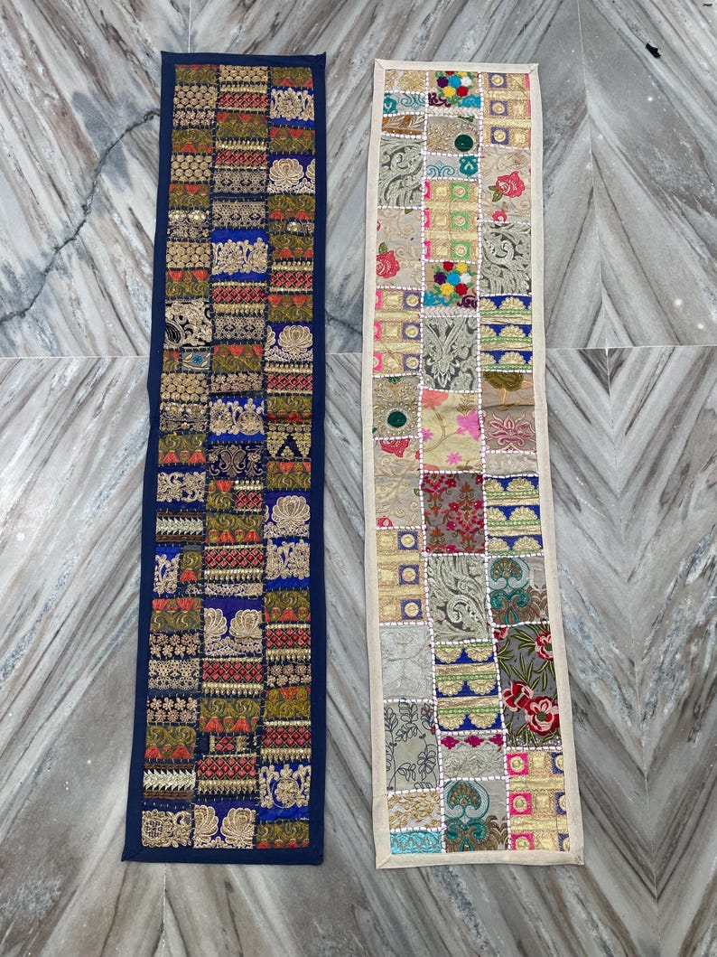 May include: Two rectangular, patchwork textile runners. One has a navy blue border and features squares of gold, red, and blue patterns. The other has an off-white border and features squares of various colors and patterns.