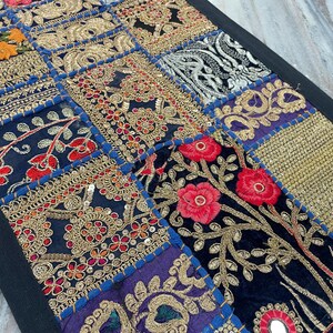 May include: A patchwork textile with a black border. The design features various embroidered squares in shades of blue, gold, red, and orange. Floral and geometric patterns are prominent, with intricate gold thread detailing and mirror accents.