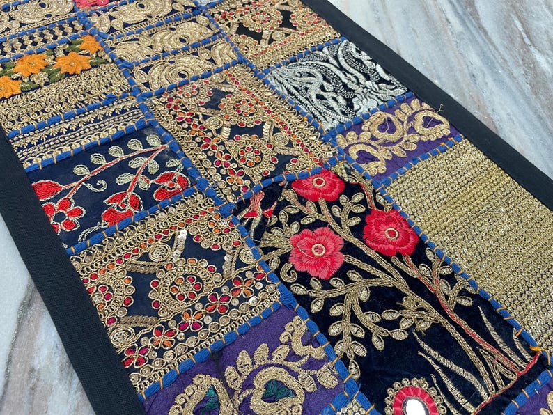 May include: A patchwork textile with a black border. The design features various embroidered squares in shades of blue, gold, red, and orange. Floral and geometric patterns are prominent, with intricate gold thread detailing and mirror accents.