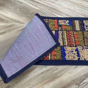May include: A rectangular, patchwork textile with a navy blue border. The patchwork features squares of gold, red, and blue fabrics with intricate embroidered designs. The reverse side is a solid lavender fabric.