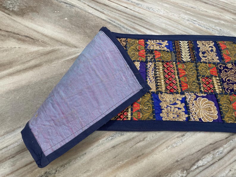 May include: A rectangular, patchwork textile with a navy blue border. The patchwork features squares of gold, red, and blue fabrics with intricate embroidered designs. The reverse side is a solid lavender fabric.