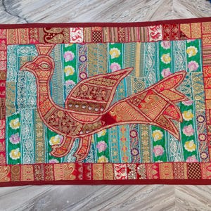 May include: A decorative textile wall hanging featuring a bird design. The bird is primarily red and gold, set against a turquoise and patterned background. The border is a deep red, and the piece has an overall patchwork aesthetic.