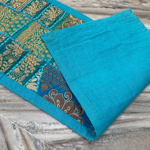 May include: A turquoise and gold embroidered table runner with a matching placemat. The runner features a patchwork design with intricate floral patterns. The placemat is a solid turquoise color, creating a complementary contrast.