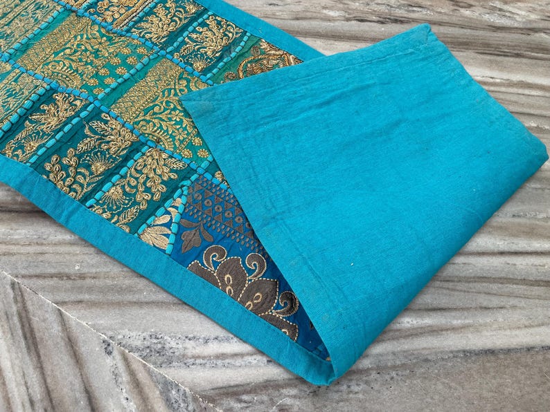 May include: A turquoise and gold embroidered table runner with a matching placemat. The runner features a patchwork design with intricate floral patterns. The placemat is a solid turquoise color, creating a complementary contrast.
