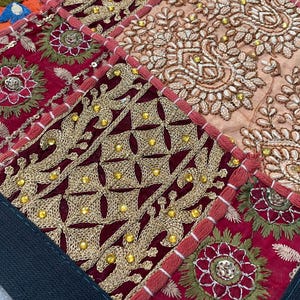 May include: Close-up of a patchwork textile with intricate embroidery. The design features squares of deep red, gold, and peach fabrics. Gold thread and small beads create detailed floral and geometric patterns. The edges are trimmed with red stitching.