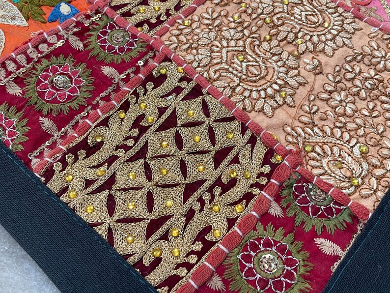 May include: Close-up of a patchwork textile with intricate embroidery. The design features squares of deep red, gold, and peach fabrics. Gold thread and small beads create detailed floral and geometric patterns. The edges are trimmed with red stitching.