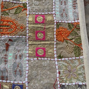 May include: Close-up of a patchwork textile with a variety of embroidered squares. The squares feature gold, pink, and neutral tones with floral and geometric patterns. Red stitching outlines each square, creating a textured, decorative piece.
