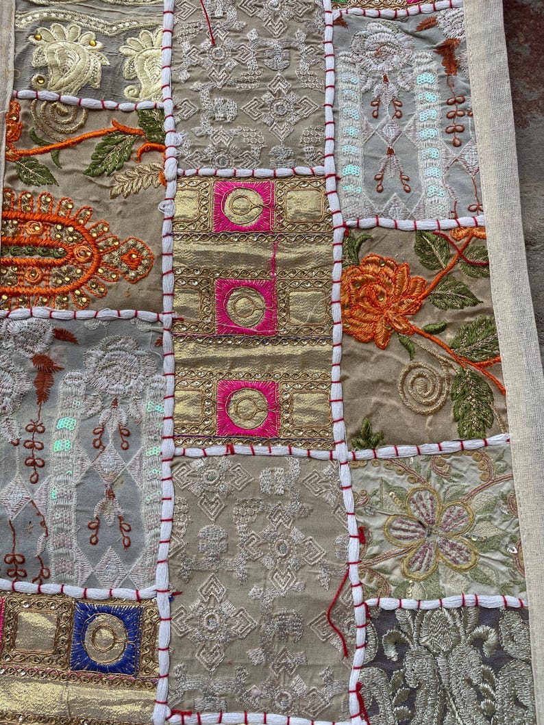 May include: Close-up of a patchwork textile with a variety of embroidered squares. The squares feature gold, pink, and neutral tones with floral and geometric patterns. Red stitching outlines each square, creating a textured, decorative piece.