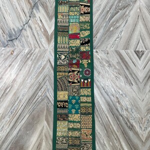 May include: A rectangular, patchwork table runner with a dark green border. The patchwork design features various patterns in shades of green, gold, red, and black. The runner is displayed on a marble surface.