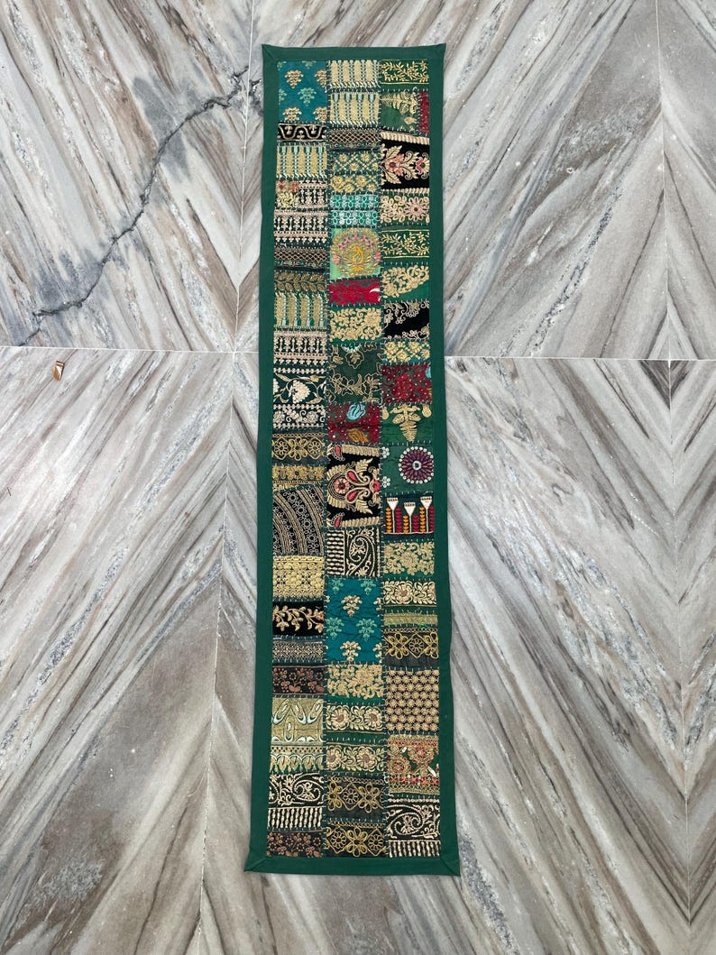 May include: A rectangular, patchwork table runner with a dark green border. The patchwork design features various patterns in shades of green, gold, red, and black. The runner is displayed on a marble surface.