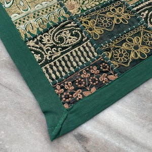 May include: Close-up of a decorative textile with a patchwork design. The textile features squares of dark green fabric with gold and bronze embroidery. A solid green border frames the patchwork, creating a cohesive look.