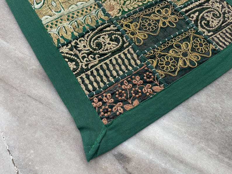 May include: Close-up of a decorative textile with a patchwork design. The textile features squares of dark green fabric with gold and bronze embroidery. A solid green border frames the patchwork, creating a cohesive look.