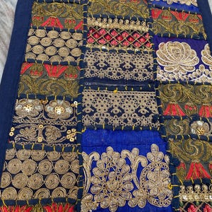 May include: A patchwork textile with a navy blue border. The design features squares of various colors, including navy blue, gold, red, and black. Intricate embroidery and sequin details adorn the fabric, creating a decorative and textured surface.