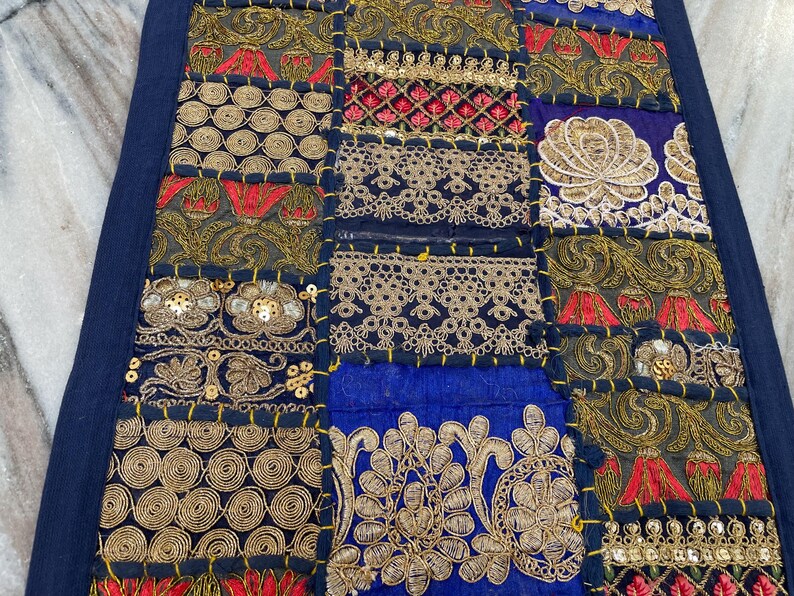 May include: A patchwork textile with a navy blue border. The design features squares of various colors, including navy blue, gold, red, and black. Intricate embroidery and sequin details adorn the fabric, creating a decorative and textured surface.