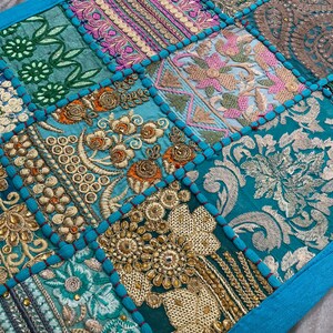 May include: A patchwork textile with a vibrant mix of colors and textures. The design features squares of teal, turquoise, and gold fabrics, adorned with intricate embroidery and beadwork. The edges are trimmed with a bright blue border, creating a decorative piece.