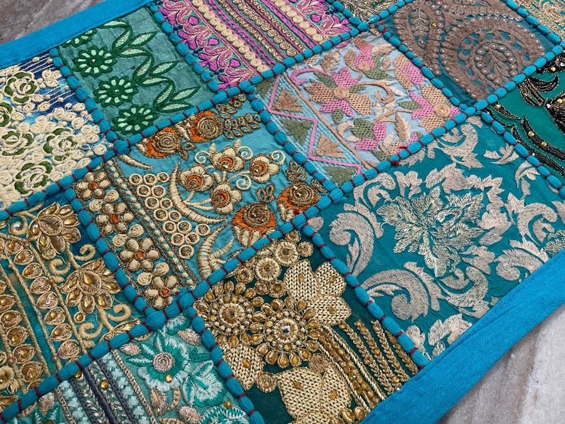 May include: A patchwork textile with a vibrant mix of colors and textures. The design features squares of teal, turquoise, and gold fabrics, adorned with intricate embroidery and beadwork. The edges are trimmed with a bright blue border, creating a decorative piece.