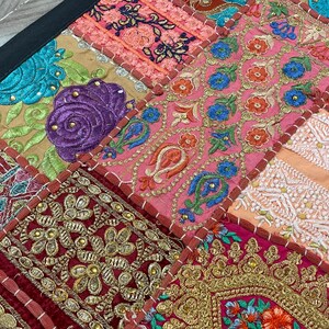 May include: Close-up of a patchwork textile with a black border. The design features squares of various colors, including pink, gold, teal, and burgundy. Each square is adorned with intricate embroidery, showcasing floral and geometric patterns. The overall aesthetic is vibrant and detailed.