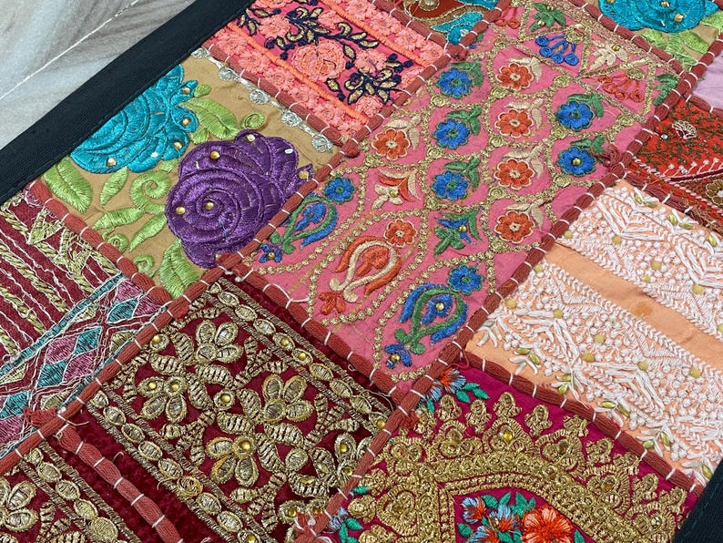 May include: Close-up of a patchwork textile with a black border. The design features squares of various colors, including pink, gold, teal, and burgundy. Each square is adorned with intricate embroidery, showcasing floral and geometric patterns. The overall aesthetic is vibrant and detailed.