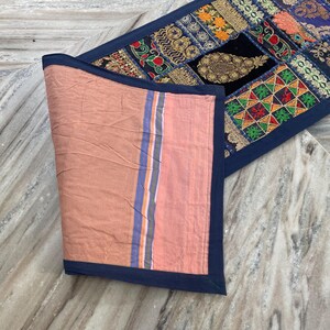 May include: A decorative table runner with a patchwork design. The runner features a variety of colorful fabric patches with intricate patterns and embroidery. The reverse side is a solid peach color with blue and orange stripes.