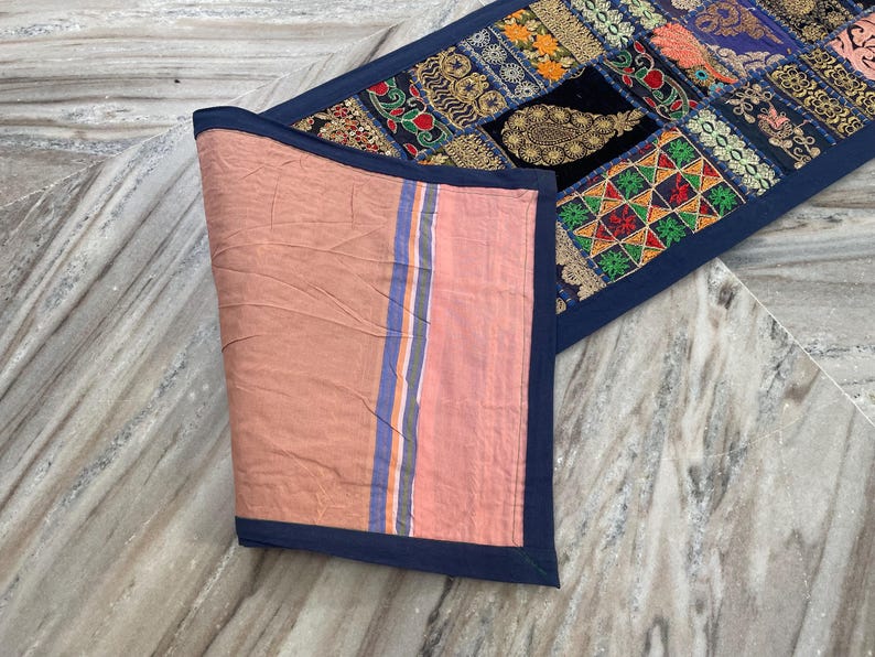 May include: A decorative table runner with a patchwork design. The runner features a variety of colorful fabric patches with intricate patterns and embroidery. The reverse side is a solid peach color with blue and orange stripes.