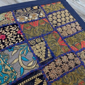May include: A patchwork textile with a dark blue border. The design features various embroidered squares with gold, red, and teal thread on a black and gray background. The embroidery includes floral and geometric patterns.