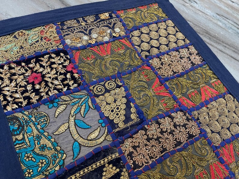 May include: A patchwork textile with a dark blue border. The design features various embroidered squares with gold, red, and teal thread on a black and gray background. The embroidery includes floral and geometric patterns.