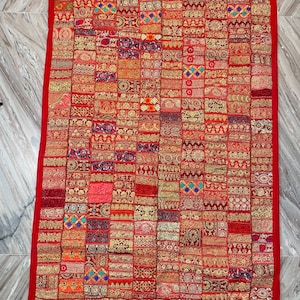 May include: A rectangular, red textile with a patchwork design. The patchwork is composed of numerous small, rectangular and square pieces in shades of red, gold, and other colors. The edges are trimmed with a solid red border. The textile is likely a decorative item.