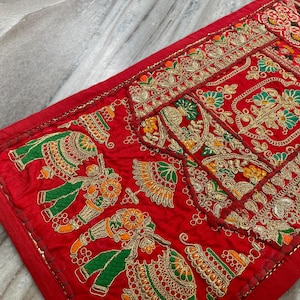 Könnte beinhalten: Ein rotes Textil mit aufwendiger Stickerei und Patchwork. Das Design zeigt detaillierte Elefantenmotive in Grün, Orange und Gold sowie florale Muster. Die Kanten sind mit Goldfaden verziert, wodurch ein dekoratives Stück entsteht.