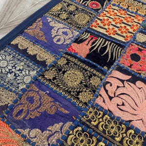 May include: A patchwork textile featuring a variety of embroidered squares in shades of blue, gold, red, and pink. The squares are bordered with a dark blue fabric. The embroidery includes floral and geometric patterns.