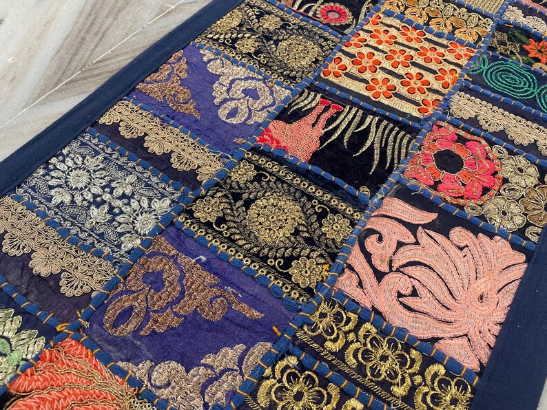 May include: A patchwork textile featuring a variety of embroidered squares in shades of blue, gold, red, and pink. The squares are bordered with a dark blue fabric. The embroidery includes floral and geometric patterns.