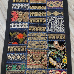 May include: A rectangular patchwork textile with a black border. The design features a variety of embroidered squares in navy blue, black, and gold, with floral and geometric patterns. The textile is likely a decorative item, such as a table runner or wall hanging.