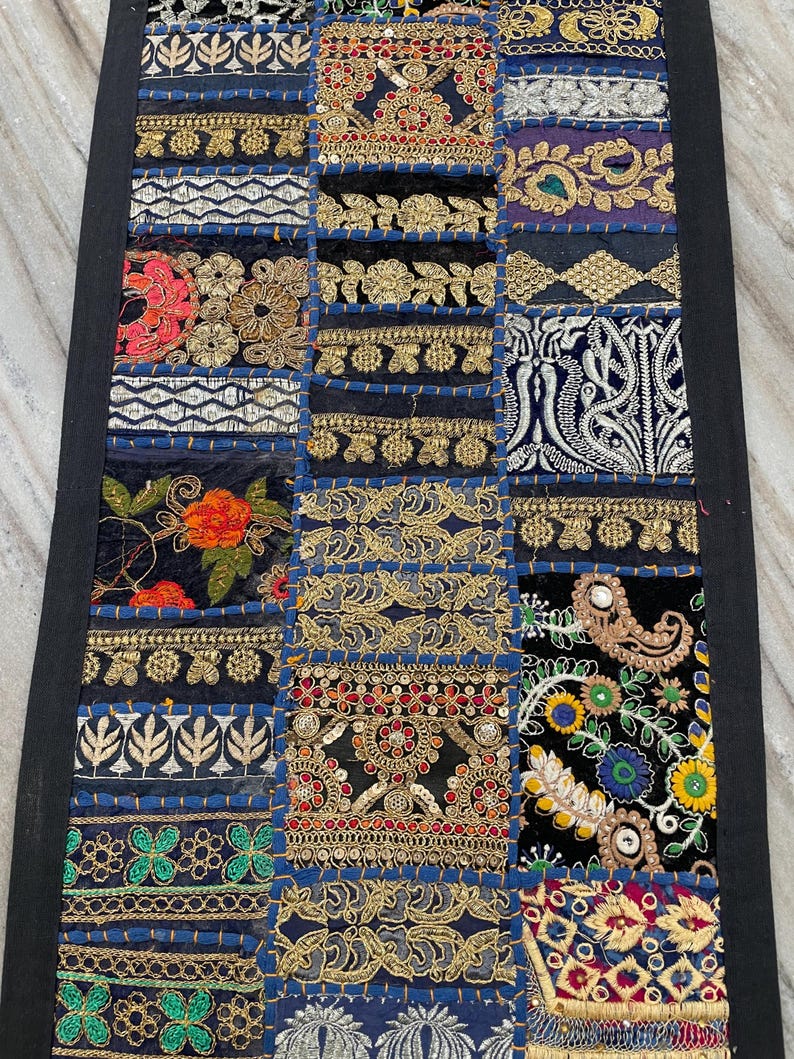 May include: A rectangular patchwork textile with a black border. The design features a variety of embroidered squares in navy blue, black, and gold, with floral and geometric patterns. The textile is likely a decorative item, such as a table runner or wall hanging.