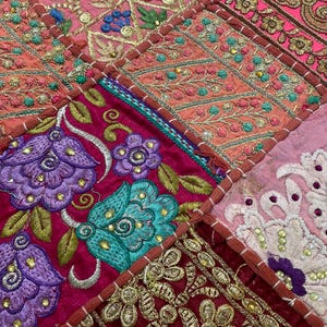 May include: Close-up of a patchwork textile with intricate embroidery. The design features squares of various colors, including pink, red, and purple, with floral and geometric patterns. The embroidery uses gold, teal, and purple threads, creating a rich, textured appearance.
