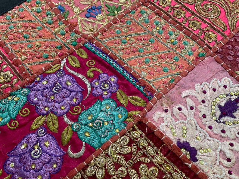 May include: Close-up of a patchwork textile with intricate embroidery. The design features squares of various colors, including pink, red, and purple, with floral and geometric patterns. The embroidery uses gold, teal, and purple threads, creating a rich, textured appearance.