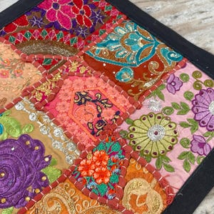 May include: A colorful, patchwork textile with a black border. The square design features embroidered squares in shades of pink, orange, gold, green, and blue. Floral and geometric patterns are visible in the detailed stitching.