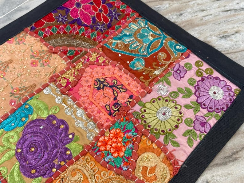 May include: A colorful, patchwork textile with a black border. The square design features embroidered squares in shades of pink, orange, gold, green, and blue. Floral and geometric patterns are visible in the detailed stitching.
