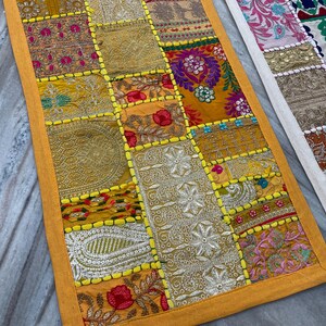 May include: A patchwork table runner with a vibrant mix of colors and patterns. The runner features squares of fabric in shades of gold, orange, and red, with intricate embroidery and embellishments. The edges are trimmed with a bright yellow border.