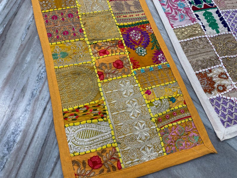 May include: A patchwork table runner with a vibrant mix of colors and patterns. The runner features squares of fabric in shades of gold, orange, and red, with intricate embroidery and embellishments. The edges are trimmed with a bright yellow border.