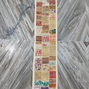 May include: A rectangular patchwork table runner with a cream-colored border. The runner is composed of various fabric squares in shades of beige, red, green, and blue. The squares feature floral and geometric patterns. The runner is placed on a marble surface.