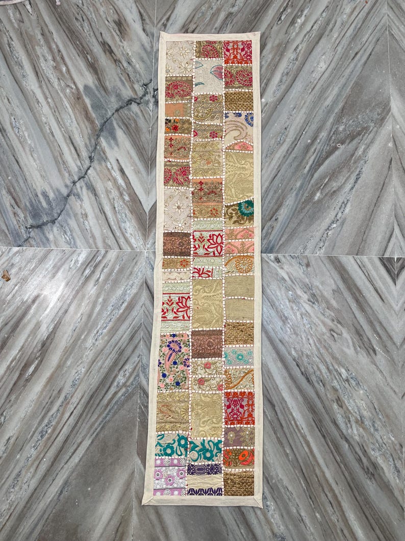 May include: A rectangular patchwork table runner with a cream-colored border. The runner is composed of various fabric squares in shades of beige, red, green, and blue. The squares feature floral and geometric patterns. The runner is placed on a marble surface.