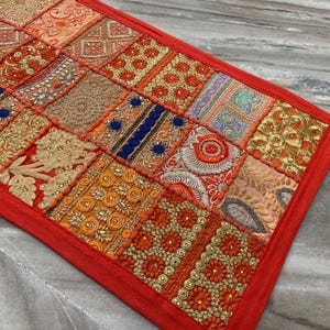 May include: A rectangular patchwork textile in shades of orange, red, gold, and blue. The design is composed of embroidered and patterned squares, framed by a solid red border. A red tassel is attached to one corner of the textile.