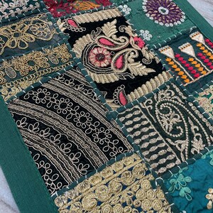 May include: A patchwork textile featuring a variety of embroidered squares in shades of green, black, and gold. The design includes floral and paisley patterns, with intricate stitching and a dark green border. The textile is likely a decorative item.
