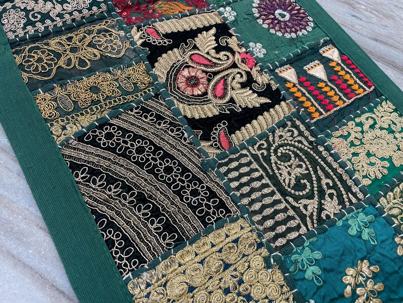 May include: A patchwork textile featuring a variety of embroidered squares in shades of green, black, and gold. The design includes floral and paisley patterns, with intricate stitching and a dark green border. The textile is likely a decorative item.