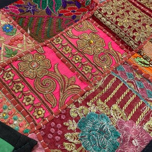 May include: Close-up of a patchwork textile with vibrant colors and intricate embroidery. The design features squares of pink, red, orange, and brown fabric, adorned with gold floral patterns and detailed stitching. The edges are finished with a black border.