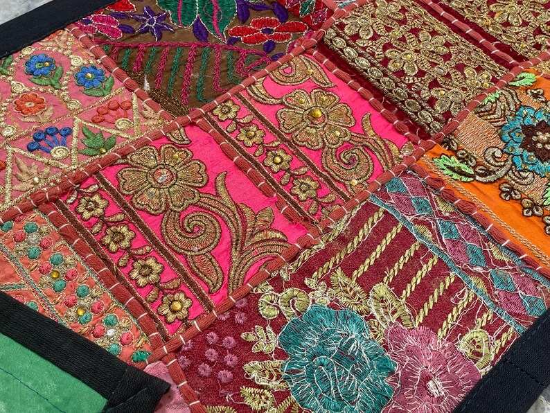May include: Close-up of a patchwork textile with vibrant colors and intricate embroidery. The design features squares of pink, red, orange, and brown fabric, adorned with gold floral patterns and detailed stitching. The edges are finished with a black border.