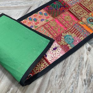 May include: A colorful patchwork table runner with a green backing and black trim. The patchwork design features various patterns and colors, including pink, orange, and gold. The runner is rectangular in shape.