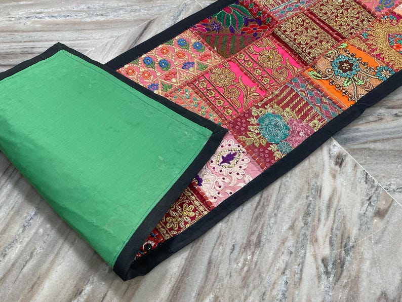 May include: A colorful patchwork table runner with a green backing and black trim. The patchwork design features various patterns and colors, including pink, orange, and gold. The runner is rectangular in shape.