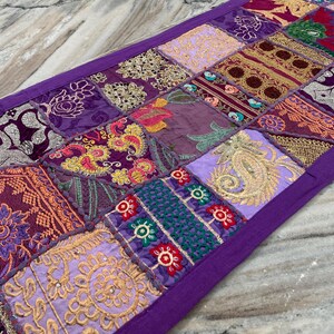 May include: A rectangular patchwork table runner with a purple border. The runner features a variety of square patches in shades of purple, gold, and burgundy, with embroidered floral and paisley designs. The runner is made of fabric.