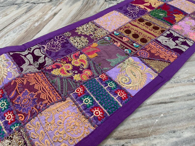 May include: A rectangular patchwork table runner with a purple border. The runner features a variety of square patches in shades of purple, gold, and burgundy, with embroidered floral and paisley designs. The runner is made of fabric.