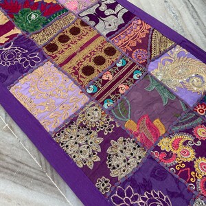 May include: A patchwork table runner with a deep purple border. The runner is composed of various fabric squares in shades of purple, gold, and pink, featuring embroidered floral and paisley designs. The runner is approximately 60 inches long.